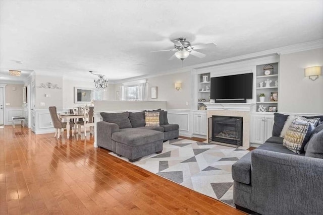 living area with built in shelves, light wood finished floors, a fireplace with flush hearth, ornamental molding, and ceiling fan with notable chandelier