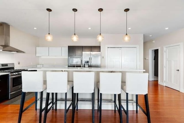 kitchen featuring stainless steel appliances, wall chimney range hood, light countertops, a breakfast bar area, and wood finished floors