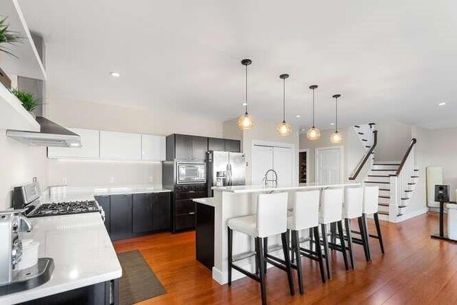 kitchen with appliances with stainless steel finishes, a breakfast bar, light countertops, wood finished floors, and wall chimney exhaust hood