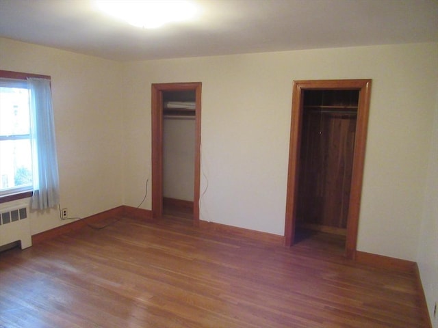 unfurnished bedroom with baseboards, two closets, wood finished floors, and radiator heating unit