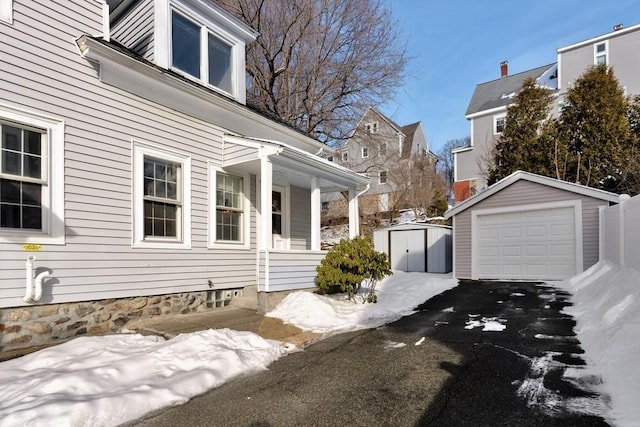 snow covered property featuring a shed, a detached garage, aphalt driveway, and an outbuilding