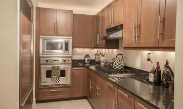 kitchen with dark stone countertops, brown cabinets, under cabinet range hood, and stainless steel appliances