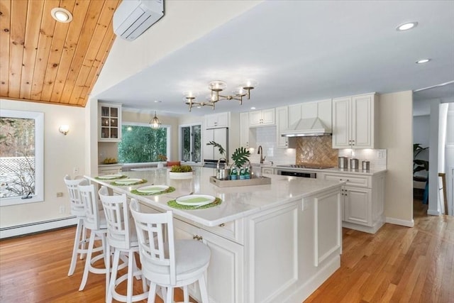 kitchen featuring a center island with sink, a baseboard radiator, custom range hood, white cabinets, and a wall mounted air conditioner