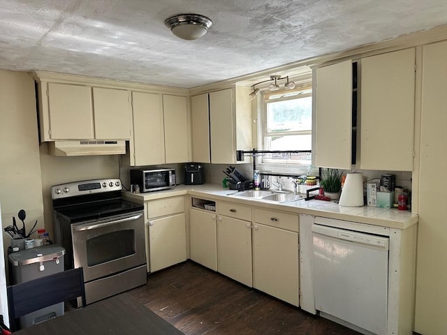 kitchen with a sink, stainless steel appliances, under cabinet range hood, cream cabinetry, and dark wood-style floors