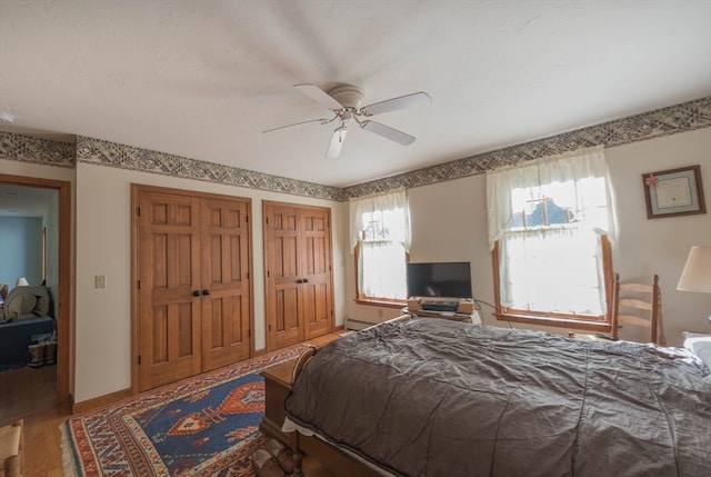 bedroom featuring ceiling fan, multiple closets, hardwood / wood-style floors, and a baseboard heating unit