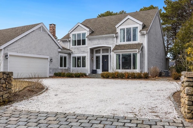 shingle-style home featuring a garage, central AC, a shingled roof, driveway, and a chimney