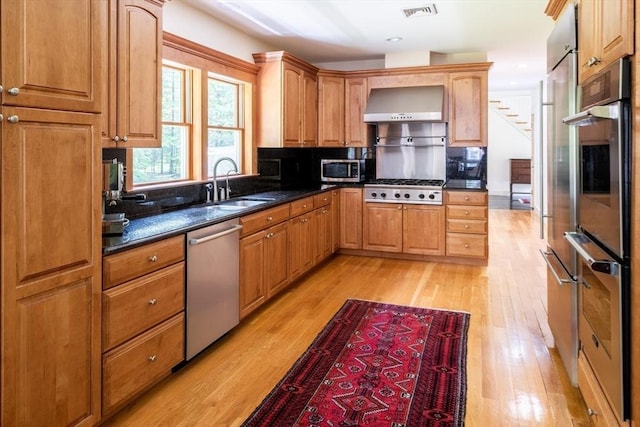 kitchen with ventilation hood, stainless steel appliances, sink, light hardwood / wood-style flooring, and dark stone countertops