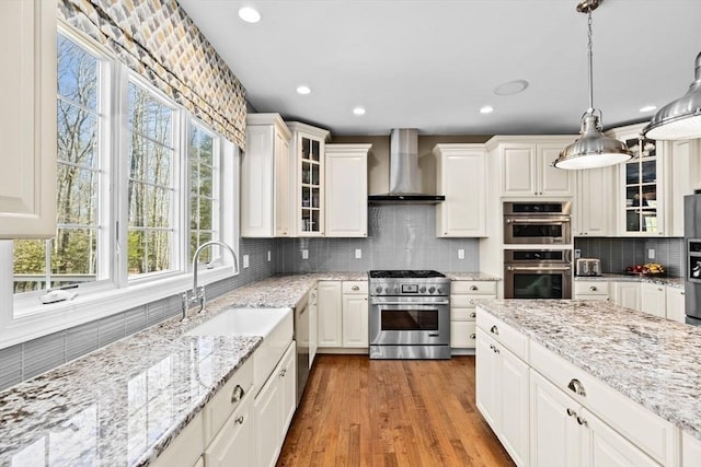 kitchen featuring a sink, wood finished floors, stainless steel appliances, wall chimney exhaust hood, and glass insert cabinets