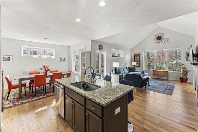 kitchen with stainless steel dishwasher, sink, light wood-type flooring, light stone countertops, and an island with sink