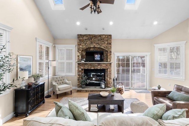 living room featuring a stone fireplace, ceiling fan, vaulted ceiling with skylight, and light hardwood / wood-style floors