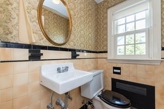 half bathroom with wainscoting, a sink, toilet, wallpapered walls, and tile walls