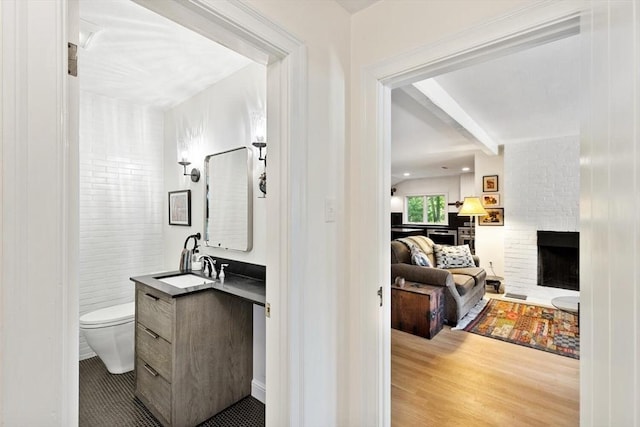 ensuite bathroom featuring a brick fireplace, beam ceiling, toilet, vanity, and wood finished floors