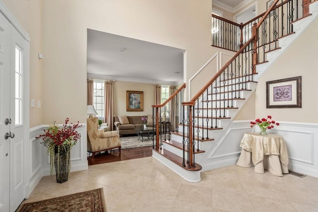 tiled entrance foyer with ornamental molding, stairs, a decorative wall, wainscoting, and a high ceiling