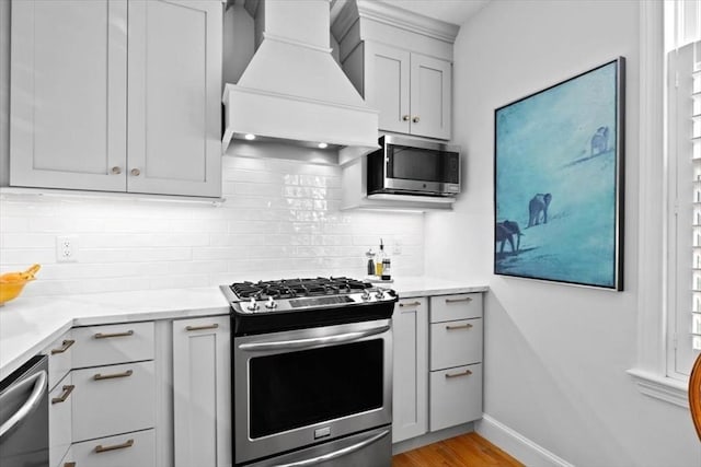 kitchen with appliances with stainless steel finishes, backsplash, light stone counters, custom exhaust hood, and light wood-type flooring