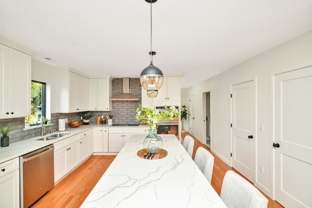 kitchen with light wood-type flooring, stainless steel dishwasher, hanging light fixtures, light stone counters, and wall chimney exhaust hood