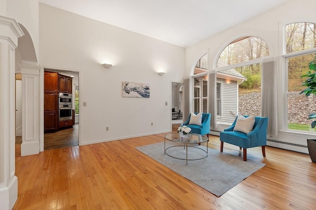 sitting room with a baseboard heating unit, arched walkways, light wood-type flooring, ornate columns, and a towering ceiling