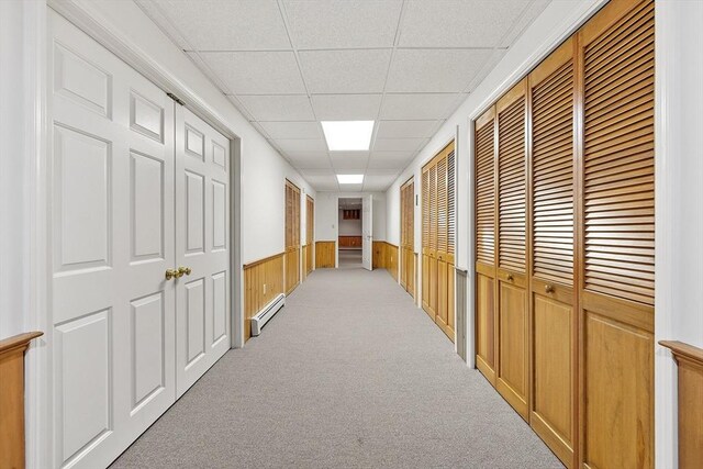 corridor featuring a baseboard heating unit, a drop ceiling, wainscoting, and light carpet