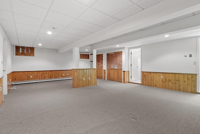 finished basement with wainscoting, light colored carpet, a baseboard radiator, a drop ceiling, and wood walls