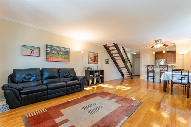 living room featuring stairway, light wood-style flooring, crown molding, and ceiling fan