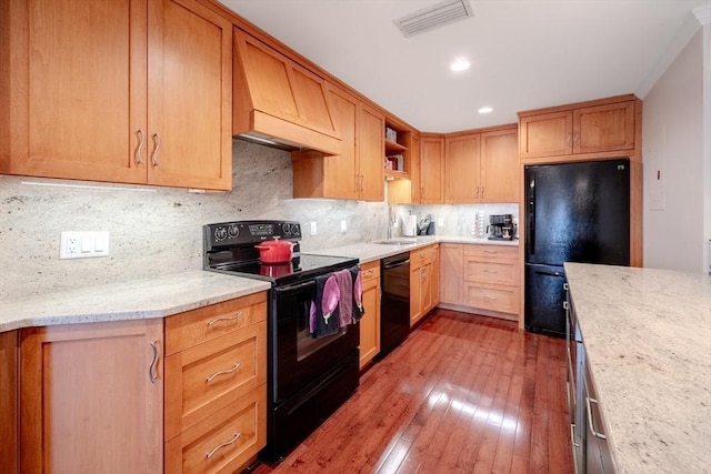 kitchen featuring tasteful backsplash, custom range hood, light stone countertops, hardwood / wood-style floors, and black appliances