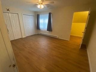 unfurnished bedroom featuring multiple closets, a ceiling fan, a baseboard heating unit, and wood finished floors