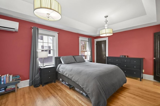 bedroom featuring light wood-type flooring, a tray ceiling, baseboards, and a wall unit AC