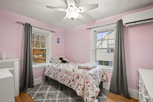 bedroom featuring a wall unit AC, wood finished floors, baseboards, and ceiling fan