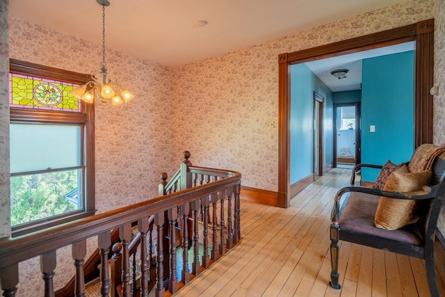 hallway featuring light wood-style floors, baseboards, wallpapered walls, a notable chandelier, and an upstairs landing