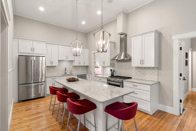 kitchen with appliances with stainless steel finishes, wall chimney range hood, ornamental molding, a breakfast bar area, and white cabinets