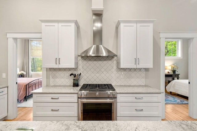 kitchen featuring stainless steel gas range, wall chimney range hood, light wood-style flooring, light stone countertops, and decorative backsplash