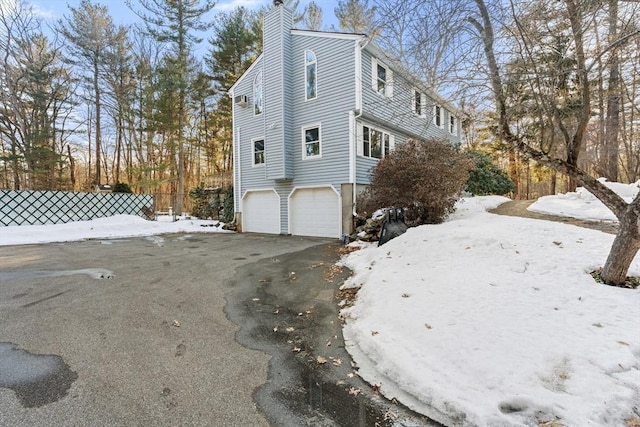 snow covered property featuring a garage, aphalt driveway, a chimney, and fence