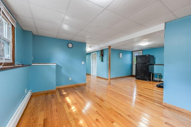 below grade area with baseboard heating, freestanding refrigerator, a drop ceiling, wood finished floors, and baseboards