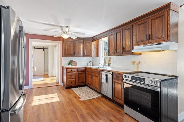 kitchen featuring light wood finished floors, appliances with stainless steel finishes, under cabinet range hood, a barn door, and tasteful backsplash