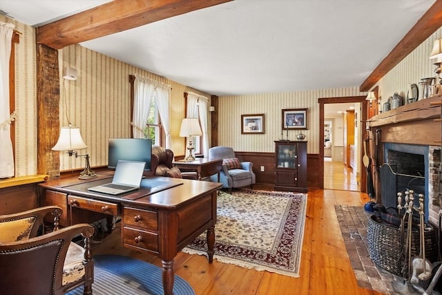 office area featuring wallpapered walls, hardwood / wood-style floors, a brick fireplace, and beam ceiling