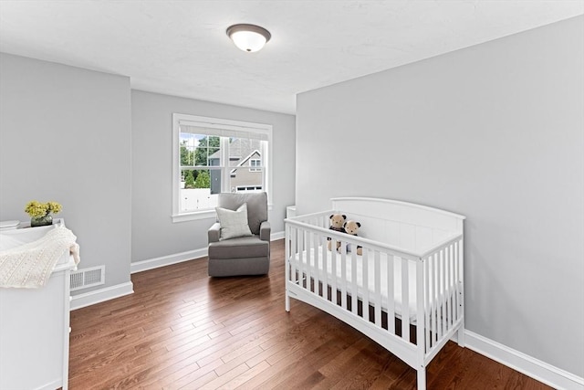 bedroom featuring a crib, dark wood finished floors, visible vents, and baseboards