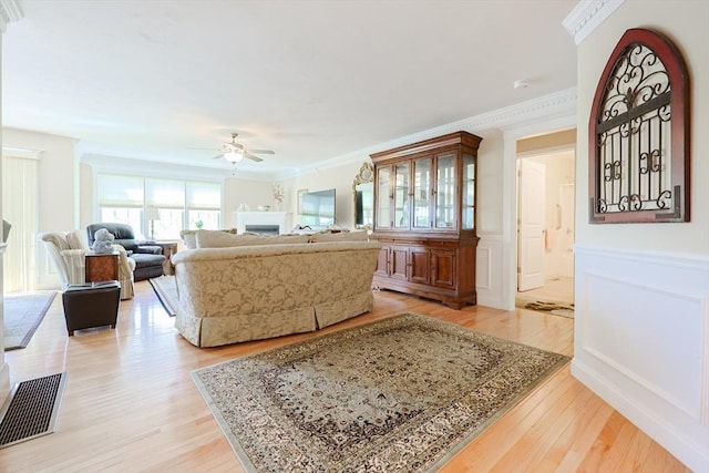 living room featuring ceiling fan, light wood-style flooring, crown molding, a fireplace, and wainscoting
