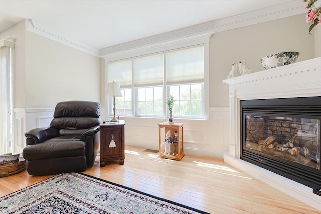 sitting room with wood-type flooring, wainscoting, a decorative wall, ornamental molding, and a glass covered fireplace