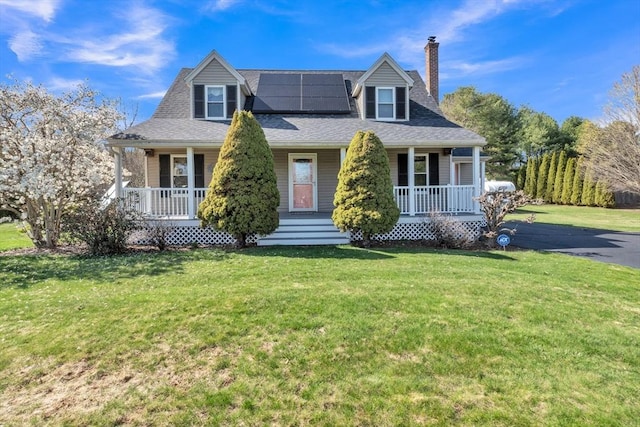 new england style home with a porch, solar panels, a chimney, roof with shingles, and a front yard