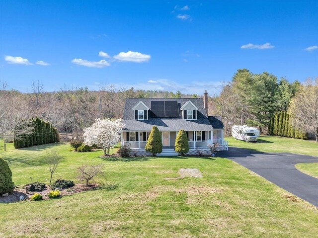 new england style home featuring a porch, driveway, a chimney, roof mounted solar panels, and a front lawn