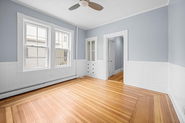 empty room with baseboard heating, wainscoting, ceiling fan, wood finished floors, and crown molding