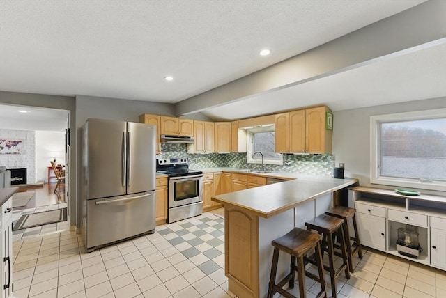 kitchen featuring tasteful backsplash, a peninsula, light brown cabinetry, under cabinet range hood, and stainless steel appliances