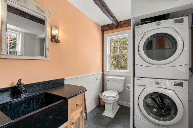 laundry room with a wainscoted wall, laundry area, dark tile patterned floors, a sink, and stacked washer / drying machine