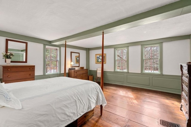 bedroom featuring a decorative wall, visible vents, wainscoting, beam ceiling, and wood-type flooring