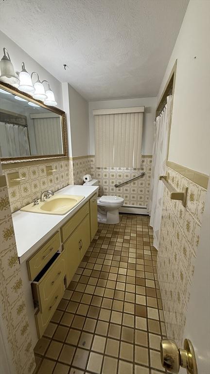 full bath with toilet, wainscoting, a textured ceiling, a baseboard radiator, and vanity