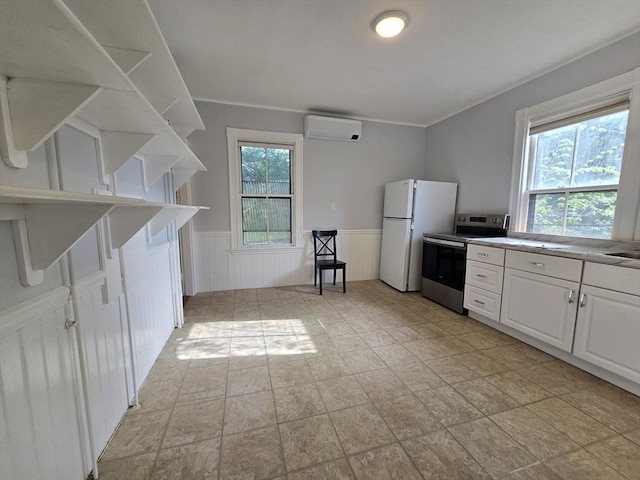 kitchen featuring stainless steel electric stove, a wall mounted air conditioner, freestanding refrigerator, a wainscoted wall, and light stone finish flooring