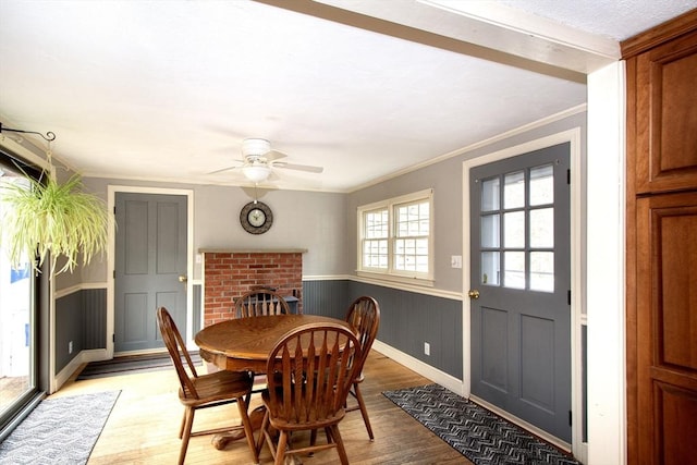 dining room with light wood finished floors, a ceiling fan, crown molding, wainscoting, and a brick fireplace