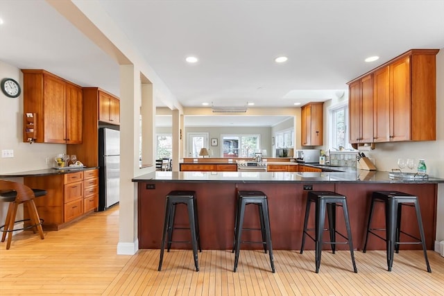 kitchen featuring light wood-style flooring, a peninsula, freestanding refrigerator, brown cabinetry, and dark countertops