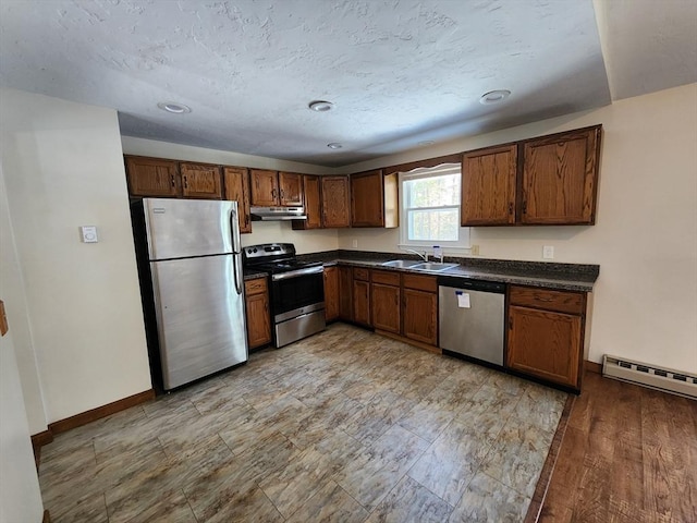 kitchen with dark countertops, a baseboard radiator, appliances with stainless steel finishes, under cabinet range hood, and a sink