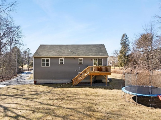 back of property with a deck, a shingled roof, stairway, a lawn, and a trampoline