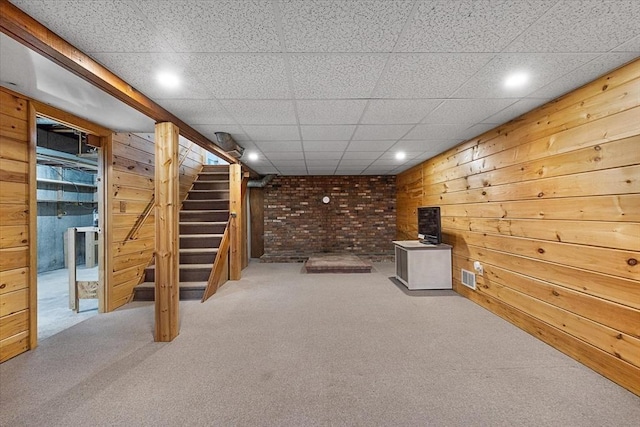 finished basement with carpet floors, visible vents, a drop ceiling, wooden walls, and stairway